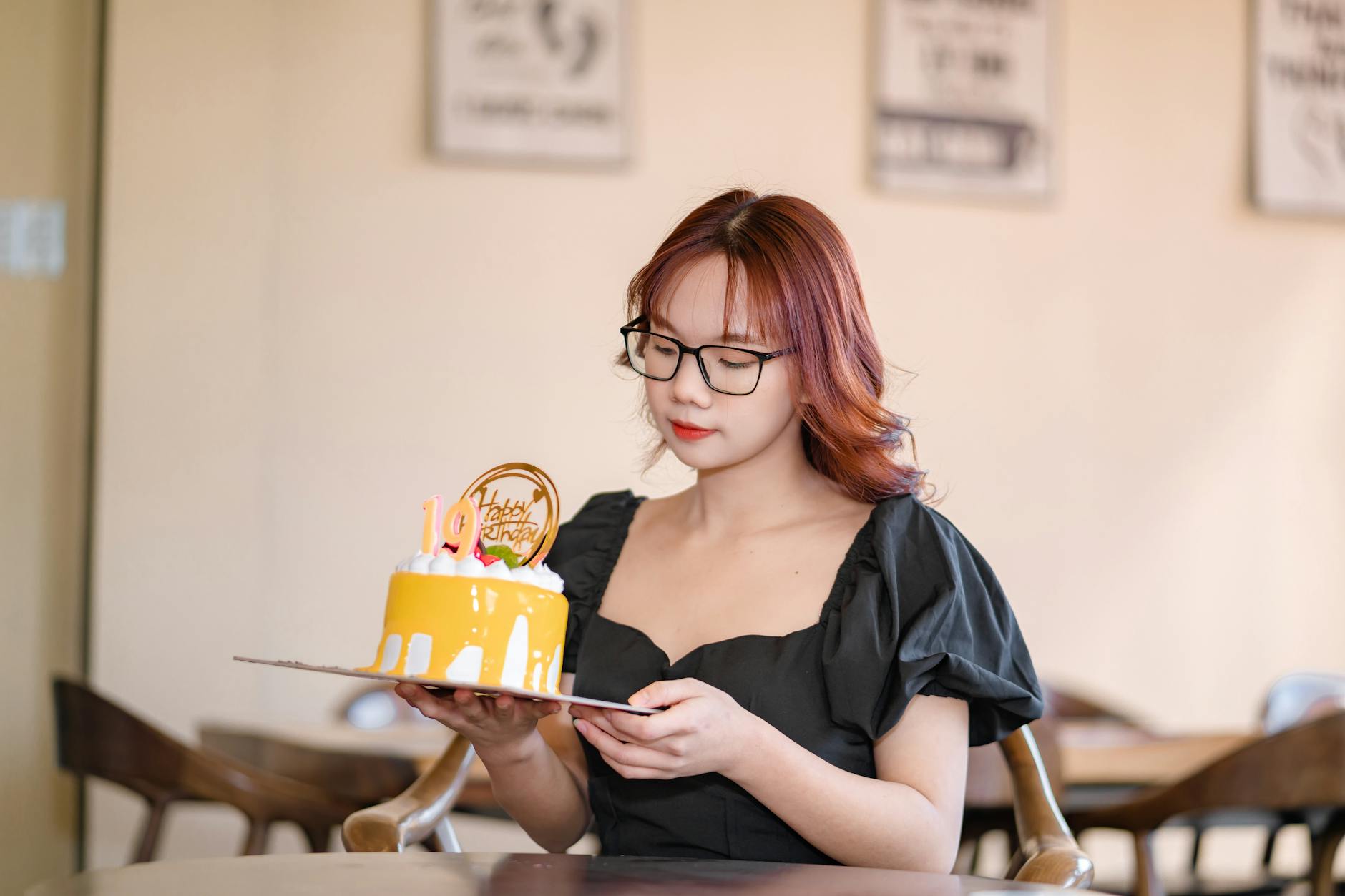 A woman in a dress holding a birthday cake with decor in a cozy cafe setting.